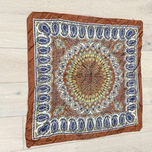 May include: Square brown scarf with a detailed paisley pattern in shades of brown, yellow, and blue. The scarf has a brown border and a central circular design. The scarf is likely made of silk or a similar fabric.