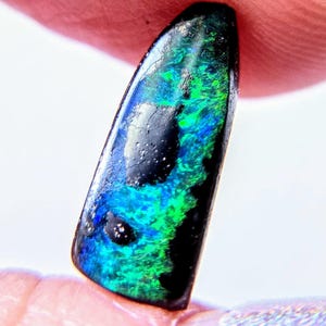 May include: A polished, elongated, teardrop-shaped black opal gemstone featuring vibrant, iridescent flashes of electric blue and bright green color play across its dark, opaque surface, showcasing a smooth, reflective, and slightly curved texture.