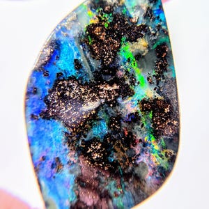 May include: A polished, teardrop-shaped boulder opal gemstone featuring a dark, ironstone matrix with vibrant flashes of electric blue, neon green, and subtle hints of red and gold throughout the iridescent, multi-colored surface.