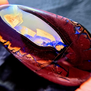 May include: An oval-shaped dark brown boulder opal gemstone featuring a prominent central vein of iridescent white, gold, and electric blue flashes. The stone displays a polished, smooth surface with deep earthy tones and vibrant colour play.