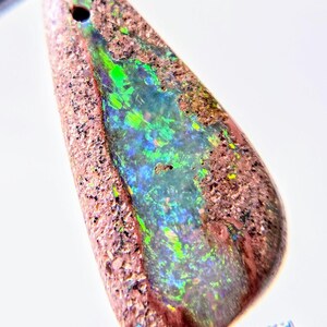 May include: A teardrop-shaped opal gemstone with a hole drilled at the top. The stone features a vibrant display of colors, including green, blue, and yellow, set against a brown matrix. The opal is held by a metal tool.