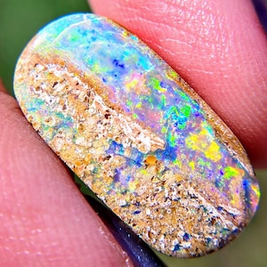 May include: An oval-shaped boulder opal gemstone featuring a polished, iridescent surface with vibrant flashes of blue, green, and yellow light. The stone displays a natural, textured tan matrix running horizontally across the center.
