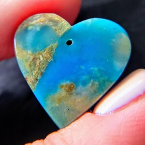 May include: A heart-shaped pendant made of polished blue stone with natural tan and brown mineral inclusions. The surface is smooth and features a small, circular hole drilled near the top center for stringing, showcasing a vibrant turquoise hue.