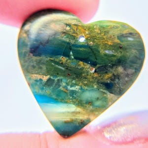 May include: A heart-shaped pendant made of polished stone featuring a variegated pattern of deep forest green, teal, and earthy brown tones with small flecks of gold. The surface is smooth and glossy, with a single small circular hole at the top.