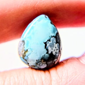 May include: A teardrop-shaped gemstone with a light blue surface and dark gray and black inclusions. The polished stone is held between two fingers, showcasing its natural patterns and color variations.