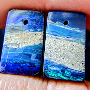 Op de afbeelding: Twee rechthoekige, gepolijste opaalhangers. Elke hanger heeft een levendige mix van blauw, groen en zilver, die doet denken aan een zeegezicht. Er zit een klein gaatje aan de bovenkant, wat duidt op sieraden.