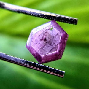 May include: A hexagonal, translucent purple gemstone held by metal tweezers. The stone features a faceted surface with visible internal inclusions and a small, circular drill hole near the top edge, showcasing a deep magenta hue throughout.