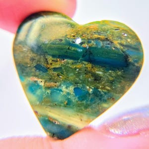 May include: A heart-shaped pendant featuring a polished surface with a marbled pattern of deep teal, forest green, and golden-brown inclusions. The stone has a smooth, glossy finish and a small, circular hole drilled near the top for suspension.
