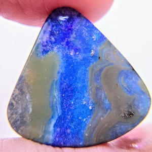 May include: A triangular-shaped gemstone with a vibrant display of colors. The stone features a deep blue hue dominating the center, complemented by swirls of gray, white, and olive green. The gemstone is held between two fingers.