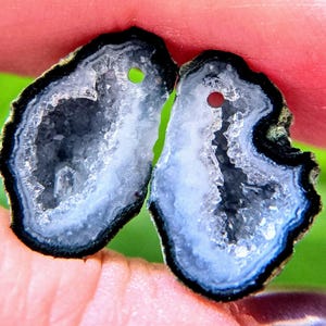 May include: Two irregular, flat agate slices featuring a dark black outer rim, a white middle band, and a sparkling, crystalline druzy center. Each stone slice has a small, circular hole drilled near the top edge for use as a pendant.