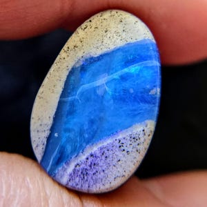 Op de afbeelding: Een gepolijste, ovale edelsteen met een opvallend blauw centrum, begrensd door witte en paarse banden. De steen heeft een gespikkelde textuur aan de randen, wat een unieke visuele aantrekkingskracht creëert. De edelsteen wordt tussen vingers gehouden.