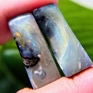 May include: Two rectangular polished gemstones featuring a smooth surface with swirling patterns of dark blue, grey, and translucent white. The stones display iridescent flashes of bright blue and yellow, with a distinct black inclusion on the left piece.