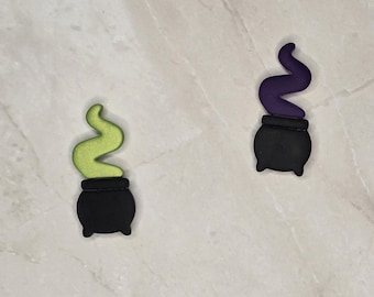 May include: Two Halloween-themed charms. One features a black cauldron with green smoke, the other a black cauldron with purple smoke. The charms are set against a light-colored, textured background.