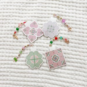 May include: Four decorative ornaments with colourful beaded accents and charms. Two ornaments feature a floral design in pink and green, while the other two have a square and a flower shape with a similar pattern. The ornaments are on a white textured surface.