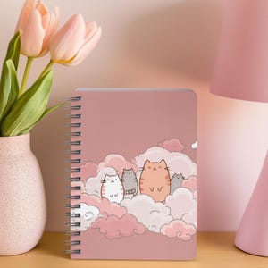 May include: A pink spiral-bound notebook with a cartoon cat illustration on the cover. The notebook is on a wooden surface, next to a vase of pink tulips and a pink lamp. The notebook's design features cats in clouds.