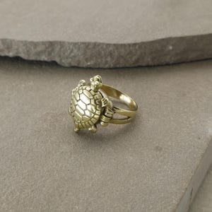 May include: A gold-colored ring featuring a detailed turtle design. The turtle has a textured shell and is positioned on the ring's band. The ring is made of a shiny, metallic material and is set against a neutral background.