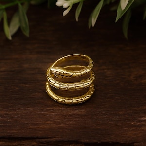 May include: A gold-colored ring in the shape of a coiled snake. The ring has three loops and a detailed snake head. The ring is on a dark wooden surface, with green and white foliage in the background.