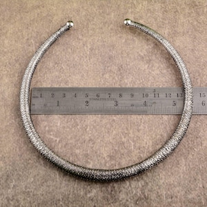 May include: Silver-toned choker necklace with a textured, scale-like pattern. The necklace is approximately 5 inches in diameter. The ends of the necklace have small, rounded ball accents. A stainless steel ruler is placed next to the necklace.