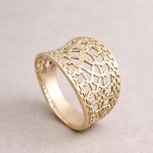 May include: A gold-colored ring with an openwork design. The ring features a wide band with a repeating pattern of geometric shapes and floral elements. The ring is displayed on a neutral background, highlighting its intricate details and craftsmanship.
