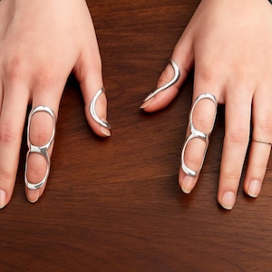 May include: Silver rings in different styles. One ring has a double-loop design, others have curved or linear shapes. The rings are shown on a dark wooden surface.