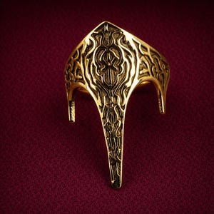 May include: A gold-colored ring with an ornate, engraved design. The ring features a central, pointed element and curved side supports. The intricate patterns and polished finish create a regal appearance. The ring is set against a deep red background.