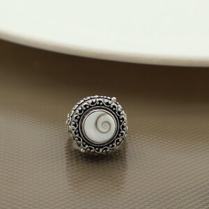 Peut inclure: Une bague en argent avec un motif en spirale blanc serti dans un cadre rond et orné. La bague présente une bordure détaillée et à motifs autour du motif central. C'est un bijou.