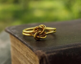 May include: A gold ring featuring a knot design, set against a blurred green backdrop. The ring sits on a dark brown, leather-bound book. The knot design is the central element, with the ring's gold colour catching the light.