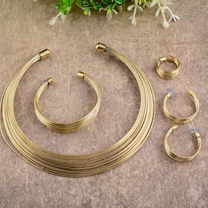 Puede incluir: Un conjunto de joyas doradas que incluye un collar de múltiples hilos, una pulsera, un anillo y pendientes de aro. El conjunto tiene un diseño minimalista con una apariencia delicada, similar a un alambre. Las joyas se exhiben sobre una superficie neutra.