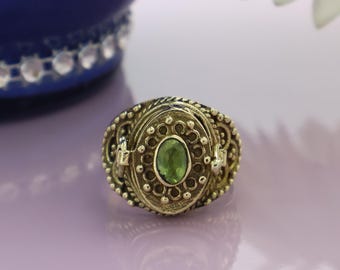 May include: A gold-toned ring with an oval green gemstone at the center. The ring features intricate filigree and beaded detailing around the gemstone and the band. The ring has a hinged compartment.