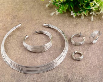 Puede incluir: Un conjunto de joyas plateadas que incluye un collar gargantilla de múltiples hilos, una pulsera a juego, pendientes de aro y un anillo. Las joyas están hechas de finos hilos de metal flexibles. El conjunto se exhibe sobre una superficie texturizada de tono neutro.