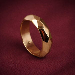 May include: A gold-colored ring with a faceted design. The ring has a geometric pattern with multiple flat surfaces that reflect light. The ring is set against a dark red background.