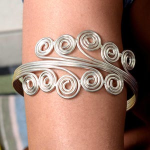 May include: Silver arm cuff with a series of spiral designs. The cuff wraps around the arm, with the spirals arranged in a flowing, decorative pattern. The metal has a bright, reflective finish, creating a striking accessory.