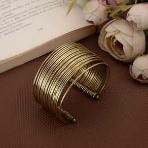 May include: A gold-toned cuff bracelet, featuring multiple parallel wire loops. The bracelet has a vintage aesthetic. It is displayed on a brown surface, with a book and flowers visible in the background.