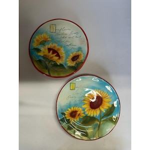 May include: Two round decorative plates with a sunflower design. The plates feature yellow sunflowers with red centres against a blue background. The plates have a red border and include script text.