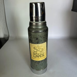 May include: A vintage green and silver thermos with a silver lid. A yellow label on the front reads "Saves Lives". The thermos has a cylindrical shape and a silver base. The lid is a shiny silver colour.