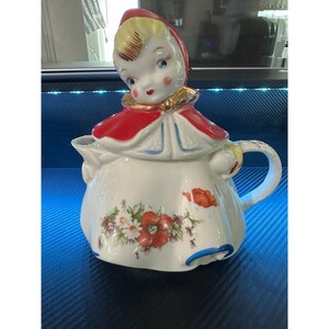 May include: A vintage ceramic teapot with a figurine of a girl in a red hooded cape. The white teapot is decorated with floral designs, including red poppies and other flowers. The handle and spout are white, with gold accents.
