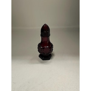 May include: A dark red glass perfume bottle with a decorative, geometric design. The bottle has a hexagonal base and a tall, pointed stopper. The body of the bottle features an embossed pattern. The overall shape is elegant and vintage.