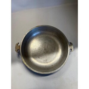May include: A round, silver-colored metal cooking pot with two curved handles on either side. The pot has a wide, shallow design and appears to be made of aluminum. The interior shows signs of use, with minor scratches and wear.