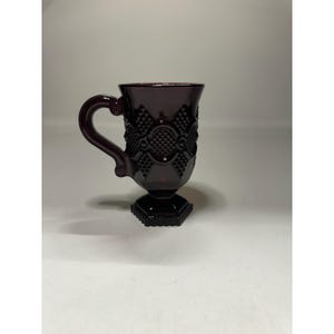 May include: A dark red glass mug with a handle and a decorative pattern. The mug has a pedestal base and a detailed design on the body and handle. The mug is likely vintage or antique.