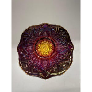 May include: A decorative, iridescent glass plate with a vibrant color gradient. The plate features a central starburst design in yellow and gold, surrounded by intricate patterns in shades of red, purple, and gold. The plate has a scalloped edge.
