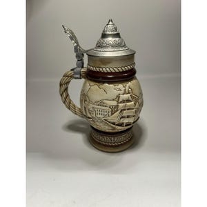 May include: A beige ceramic beer stein with a silver metal lid and handle. The stein features a nautical scene with a ship and building. The handle is rope-like, and the base has text. A decorative item.