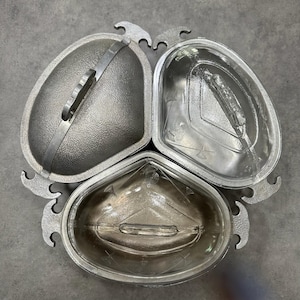 May include: A three-section serving dish with a silver finish. Each section is a different shape, with one having a lid and handle. The dish has decorative curved handles on the outside edges, suitable for serving appetizers or snacks.