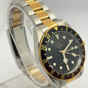 May include: A Tudor Geneve wristwatch featuring a black dial with luminous hour markers, gold-tone hands, and a date window. The watch has a two-tone stainless steel and gold bracelet and a rotating bezel with a black and burgundy 24-hour scale.