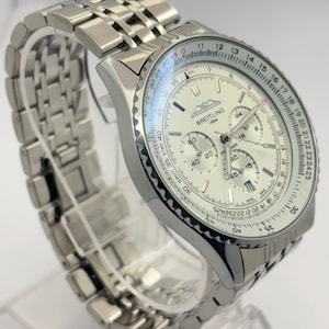 Könnte beinhalten: Eine silberfarbene Breitling 1884 Chronographenuhr mit einem Metallgliederarmband. Die Uhr verfügt über ein weißes Zifferblatt mit drei Hilfszifferblättern, einem Datumsfenster und einer drehbaren Rechenschieber-Lünette mit schwarzen Zahlen.