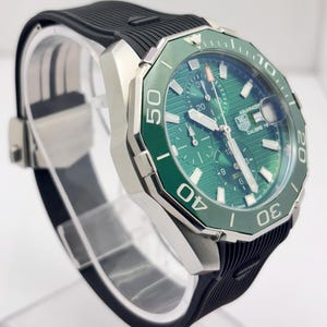 Pode incluir: Um relógio prateado com mostrador e moldura verdes e uma bracelete de borracha preta. O mostrador do relógio exibe vários mostradores e o texto "AQUARACER CALIBRE 16". A moldura tem algarismos brancos. O relógio é exibido num suporte transparente.