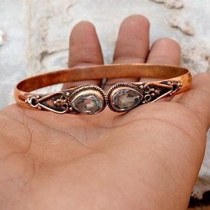 以下が含まれることがあります： 2つのペアシェイプのライトブルーの宝石が付いた銅製のカフブレスレット。ブレスレットは、装飾的なディテールを備えたオープンデザインです。金属は暖かく、赤みがかった茶色の色合いをしています。