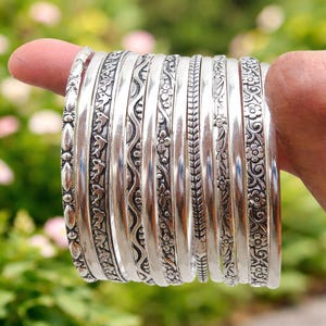 May include: A collection of silver bangles with intricate floral and geometric patterns. The bangles are held up, showcasing their detailed designs and polished finish. The bangles are a mix of different patterns, adding visual interest.