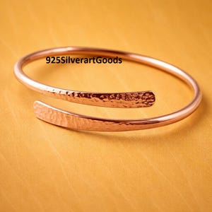 May include: A copper-coloured, open cuff bracelet with a hammered texture. The bracelet has a simple, elegant design and is set against a yellow background. The text "925SilverartGoods" is visible on the bracelet.