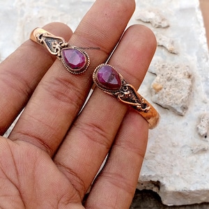 May include: Two copper-colored bracelets with ornate detailing and teardrop-shaped, deep red gemstones. The bracelets are displayed on a textured surface. The bracelets have a vintage or antique aesthetic.