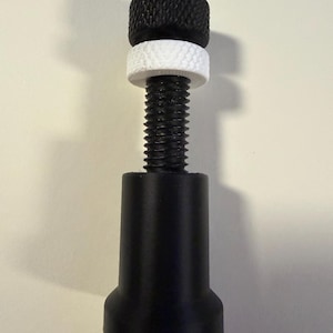May include: A black adjustable furniture leg with a textured black knob, a white ring, and a threaded rod. The base of the leg is also black. This product is designed for furniture support and leveling.